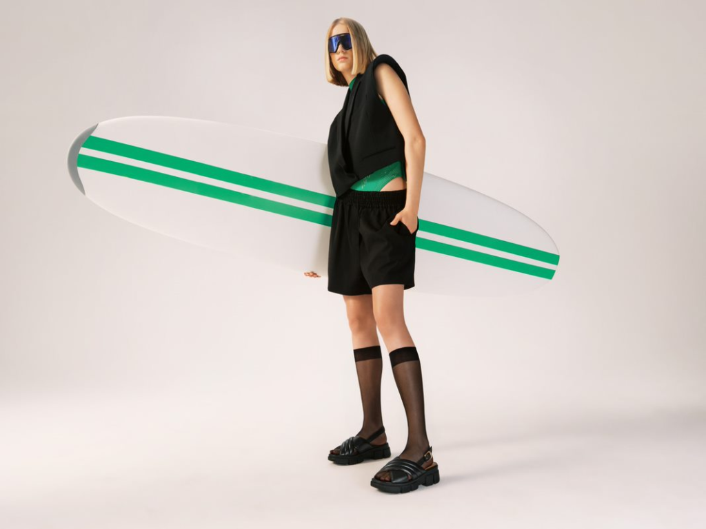 Modelka v čiernom totálnom vzhľade s čiernymi sandálmi, držiaca surfovacie prkno - kampaň na leto 2023